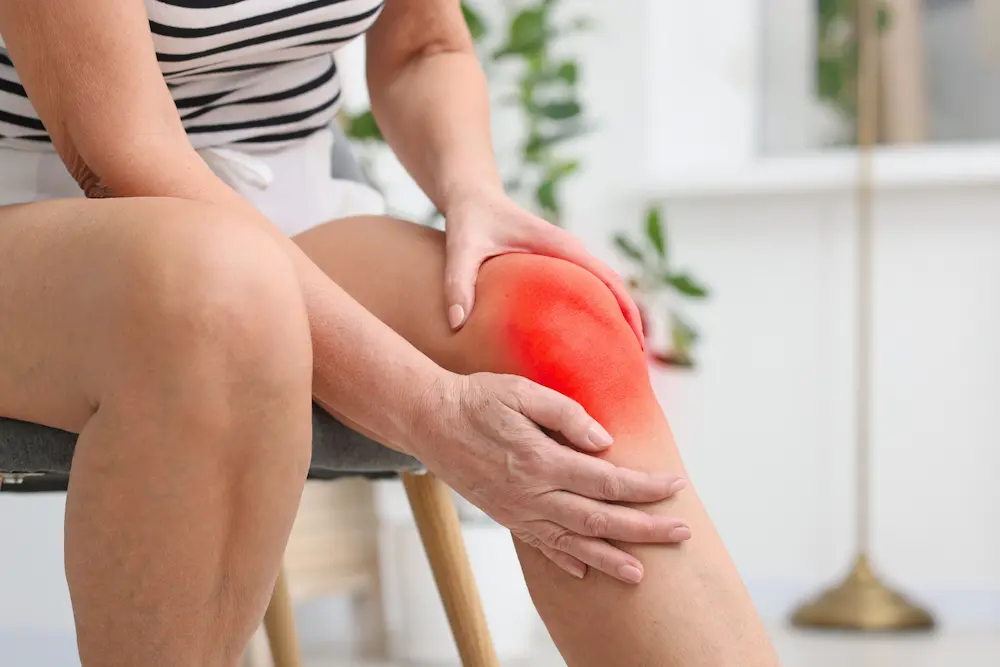 Patient at home experiencing knee pain sitting on a chair with a plant in the background
